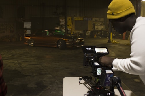 A man wearing a yellow beanie and a white shirt is operating a professional camera setup on a tripod, capturing footage of a brown, modified car performing a drift inside a warehouse with dim lighting. The warehouse has various signs and automotive equipment in the background.