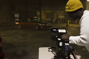 A man wearing a yellow beanie and a white shirt is operating a professional camera setup on a tripod, capturing footage of a brown, modified car performing a drift inside a warehouse with dim lighting. The warehouse has various signs and automotive equipment in the background.