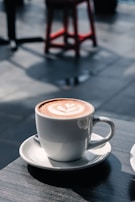 A steaming cup of coffee with latte art on its surface is placed on a saucer. The cup sits on a table with a blurred background, including a red bar stool and soft lighting creating shadows.