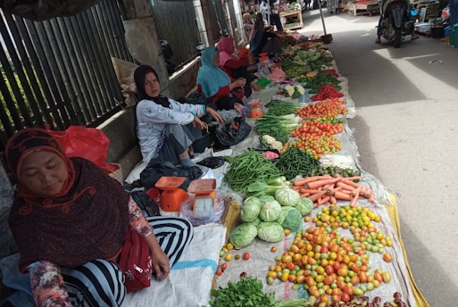 A market scene featuring several women sitting on the ground selling an array of fresh vegetables. The produce includes tomatoes, green beans, cabbages, carrots, and chilies, arranged in neat piles. The surroundings are outdoor with a metal fence visible in the background.