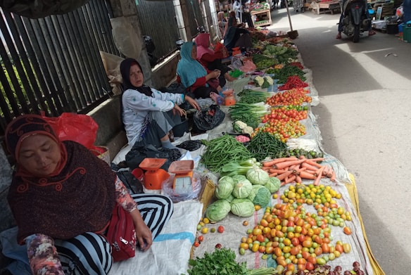 A market scene featuring several women sitting on the ground selling an array of fresh vegetables. The produce includes tomatoes, green beans, cabbages, carrots, and chilies, arranged in neat piles. The surroundings are outdoor with a metal fence visible in the background.