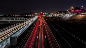 A nighttime view of a highway with blurred lights from moving vehicles creating streaks of red and white along the road. Illuminated signs for 'BOSCH' and 'L-BANK' are visible on buildings adjacent to the road. Streetlights cast a subtle glow over the scene, with distant city lights in the background.
