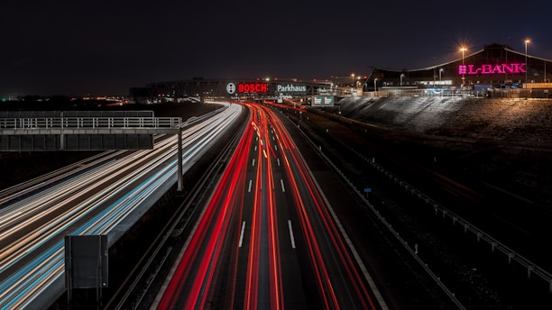 A nighttime view of a highway with blurred lights from moving vehicles creating streaks of red and white along the road. Illuminated signs for 'BOSCH' and 'L-BANK' are visible on buildings adjacent to the road. Streetlights cast a subtle glow over the scene, with distant city lights in the background.