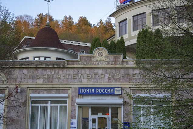 A historic building with a sign in Russian, indicating it is a post office. The architecture features a rounded structure with traditional design elements. Surrounding the building are trees and vegetation, and there is a visible sign with the name 'Post of Russia' near the entrance.