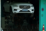 A white car with a blue license plate is elevated on a hydraulic lift inside a workshop or garage with a teal-colored wall and a safety cone nearby.