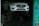 A white car with a blue license plate is elevated on a hydraulic lift inside a workshop or garage with a teal-colored wall and a safety cone nearby.