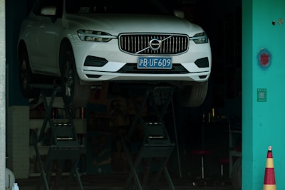 A white car with a blue license plate is elevated on a hydraulic lift inside a workshop or garage with a teal-colored wall and a safety cone nearby.