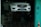 A white car with a blue license plate is elevated on a hydraulic lift inside a workshop or garage with a teal-colored wall and a safety cone nearby.