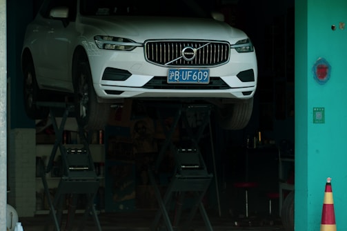 A white car with a blue license plate is elevated on a hydraulic lift inside a workshop or garage with a teal-colored wall and a safety cone nearby.