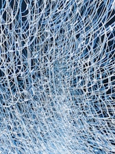 An intricate and complex network of intertwined lines and patterns, forming a mesh-like structure with a sense of movement and flow. The lines are predominantly white against a dark background, giving a sense of depth.