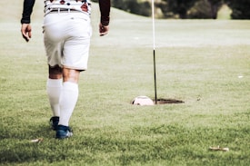A person wearing a patterned shirt, white shorts, and knee-high socks walks across a grassy area toward a large golf hole containing a football. The scene is set in an outdoor environment, likely on a golf course.