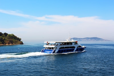 A ferry navigates through calm blue waters under a clear sky. The vessel is white with blue accents and a flag on top. In the background, an island with lush greenery and a mountain is visible, adding to the serene setting.