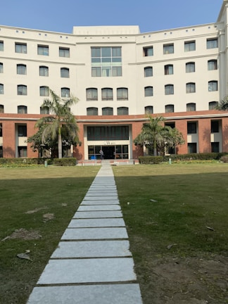 A multi-story building with a white facade and brown accents, featuring a large number of square windows. The entrance is marked by a line of palm trees and a pathway leading up to it. The foreground consists of a neatly maintained lawn.