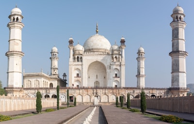 A grand white marble mausoleum with a large central dome flanked by four smaller domes. Tall minarets stand at each corner, and the architecture features intricate carvings and arches. The structure is surrounded by a landscaped garden and pathways lined with fountains and plants.