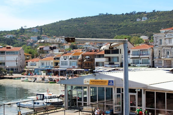 A coastal area with a pier labeled 'Burgazada Iskelesi' in front of a row of colorful buildings. The buildings have red-tiled roofs and face the water, with a green hillside in the background. Small boats are docked along the waterfront, and there is a streetlamp in the foreground.