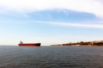 A cargo ship loaded with oil barrels sailing near the Dubai coastline under a clear sky.