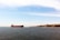 A large cargo ship is sailing in a wide expanse of calm water, with a city skyline featuring historic buildings and minarets in the background under a clear blue sky.
