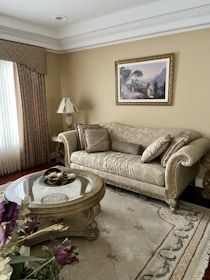 A well-decorated living room features an ornate beige sofa adorned with matching cushions, placed against a light brown wall. Above the sofa, a framed landscape painting adds classic charm. A round, glass-top coffee table with intricate designs holds decorative items. The room is completed with a patterned rug and draped curtains. A bouquet of flowers is visible in the foreground, contributing to a warm and inviting atmosphere.