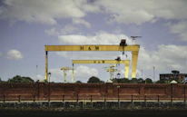 Two large, yellow construction cranes with the letters H & W are positioned behind a red brick wall. There are smaller cranes in the background, along with some trees and buildings. The sky is partly cloudy.