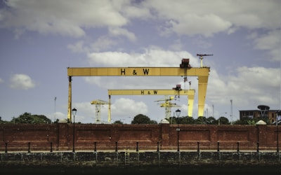 Two large, yellow construction cranes with the letters H & W are positioned behind a red brick wall. There are smaller cranes in the background, along with some trees and buildings. The sky is partly cloudy.