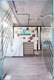 A modern entrance to a co-working space featuring glass doors with diagonal patterns, leading into a room with a decorative wall. The wall includes various objects and an eye-catching logo for the space. The floors are wooden and there are visible industrial-style ceiling fixtures.