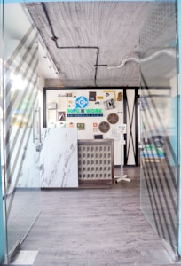 A modern entrance to a co-working space featuring glass doors with diagonal patterns, leading into a room with a decorative wall. The wall includes various objects and an eye-catching logo for the space. The floors are wooden and there are visible industrial-style ceiling fixtures.