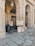 An artist is sitting on a small stool, working on a portrait painting under a stone archway. Several completed portraits are displayed on easels. The setting appears to be an outdoor, covered gallery with large stone columns and arched ceilings.