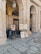 An artist is sitting on a small stool, working on a portrait painting under a stone archway. Several completed portraits are displayed on easels. The setting appears to be an outdoor, covered gallery with large stone columns and arched ceilings.