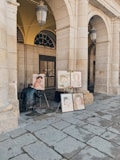 An artist is sitting on a small stool, working on a portrait painting under a stone archway. Several completed portraits are displayed on easels. The setting appears to be an outdoor, covered gallery with large stone columns and arched ceilings.