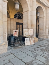 An artist is sitting on a small stool, working on a portrait painting under a stone archway. Several completed portraits are displayed on easels. The setting appears to be an outdoor, covered gallery with large stone columns and arched ceilings.