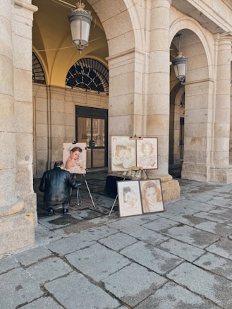 An artist is sitting on a small stool, working on a portrait painting under a stone archway. Several completed portraits are displayed on easels. The setting appears to be an outdoor, covered gallery with large stone columns and arched ceilings.