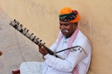 A man wearing a vibrant orange and green turban with a mustache and white shirt sits against a beige wall, playing a traditional string instrument. The instrument features multiple tuning pegs and appears handcrafted. The scene suggests an outdoor setting on a stone pavement.