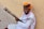 A man wearing a vibrant orange and green turban with a mustache and white shirt sits against a beige wall, playing a traditional string instrument. The instrument features multiple tuning pegs and appears handcrafted. The scene suggests an outdoor setting on a stone pavement.