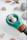 A collection of small seashells is arranged on a smooth, white surface. A wooden object featuring a turquoise segment holds two prominent seashells, one of which is spiral-shaped. Around it, more seashells and stones are scattered, creating a natural and serene composition.