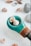 A collection of small seashells is arranged on a smooth, white surface. A wooden object featuring a turquoise segment holds two prominent seashells, one of which is spiral-shaped. Around it, more seashells and stones are scattered, creating a natural and serene composition.