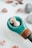 A collection of small seashells is arranged on a smooth, white surface. A wooden object featuring a turquoise segment holds two prominent seashells, one of which is spiral-shaped. Around it, more seashells and stones are scattered, creating a natural and serene composition.
