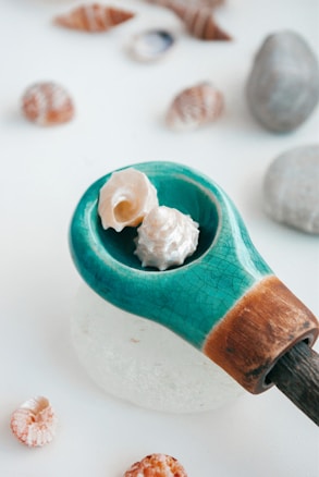 A collection of small seashells is arranged on a smooth, white surface. A wooden object featuring a turquoise segment holds two prominent seashells, one of which is spiral-shaped. Around it, more seashells and stones are scattered, creating a natural and serene composition.