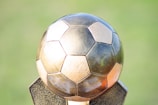 A metallic soccer ball sculpture with pentagonal and hexagonal panels on a textured base, set against a blurred green background.