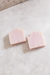 Two rectangular pieces of soap with a light pink hue rest on a smooth, light gray surface. Each soap piece has a few darker speckles scattered across its surface, and the edges appear slightly rough.