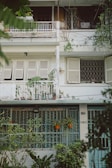 A three-story residential building facade with a vintage look, featuring shuttered windows and metal railings. There is abundant greenery with plants in pots and hanging from the balcony. The building has a slightly worn appearance with some peeling paint, and the number 004 is visible next to one of the doors.