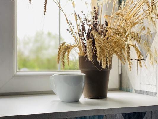 A white ceramic mug sits on a windowsill beside a potted arrangement of dried wheat and lavender. Soft natural light filters through the window, creating a calm and serene atmosphere.