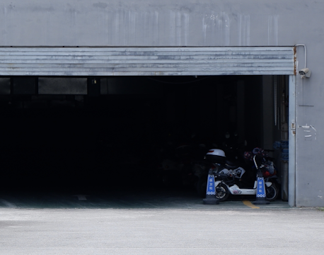 A partially open garage door reveals a dimly lit interior with several parked scooters. The entrance is bordered by traffic safety cones, and the outer wall of the garage appears worn with traces of rust near the door mechanism.