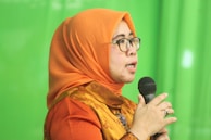 A person wearing an orange headscarf and shirt speaks into a microphone. They are wearing glasses and appear to be speaking with expression. The background is a solid green color.