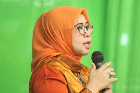 A person wearing an orange headscarf and shirt speaks into a microphone. They are wearing glasses and appear to be speaking with expression. The background is a solid green color.