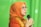 A person wearing an orange headscarf and shirt speaks into a microphone. They are wearing glasses and appear to be speaking with expression. The background is a solid green color.