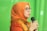 A person wearing an orange headscarf and shirt speaks into a microphone. They are wearing glasses and appear to be speaking with expression. The background is a solid green color.