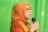 A person wearing an orange headscarf and shirt speaks into a microphone. They are wearing glasses and appear to be speaking with expression. The background is a solid green color.