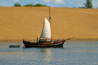 A single-masted wooden sailboat is floating on a calm body of water. The sail is partially unfurled, and there is a person on the boat, seemingly adjusting the sail or performing some task. The background features a large sandy embankment with sparse greenery at the top, under a blue sky with some clouds.