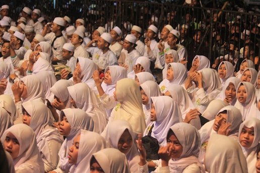 A large group of people dressed in white robes and headscarves are seated closely together, appearing to be part of a communal gathering or religious event. Many have their hands raised, possibly in prayer or singing.