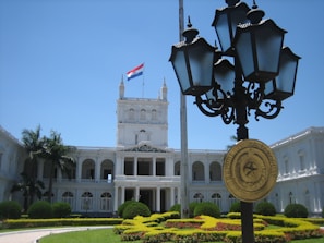 A majestic, white neoclassical building with multiple arches and columns, surrounded by lush greenery and well-maintained gardens. A flag featuring red, white, and blue stripes is flying atop the building. In the foreground, a decorative street lamp with a plaque reading 'Republica del Paraguay' is prominently displayed.